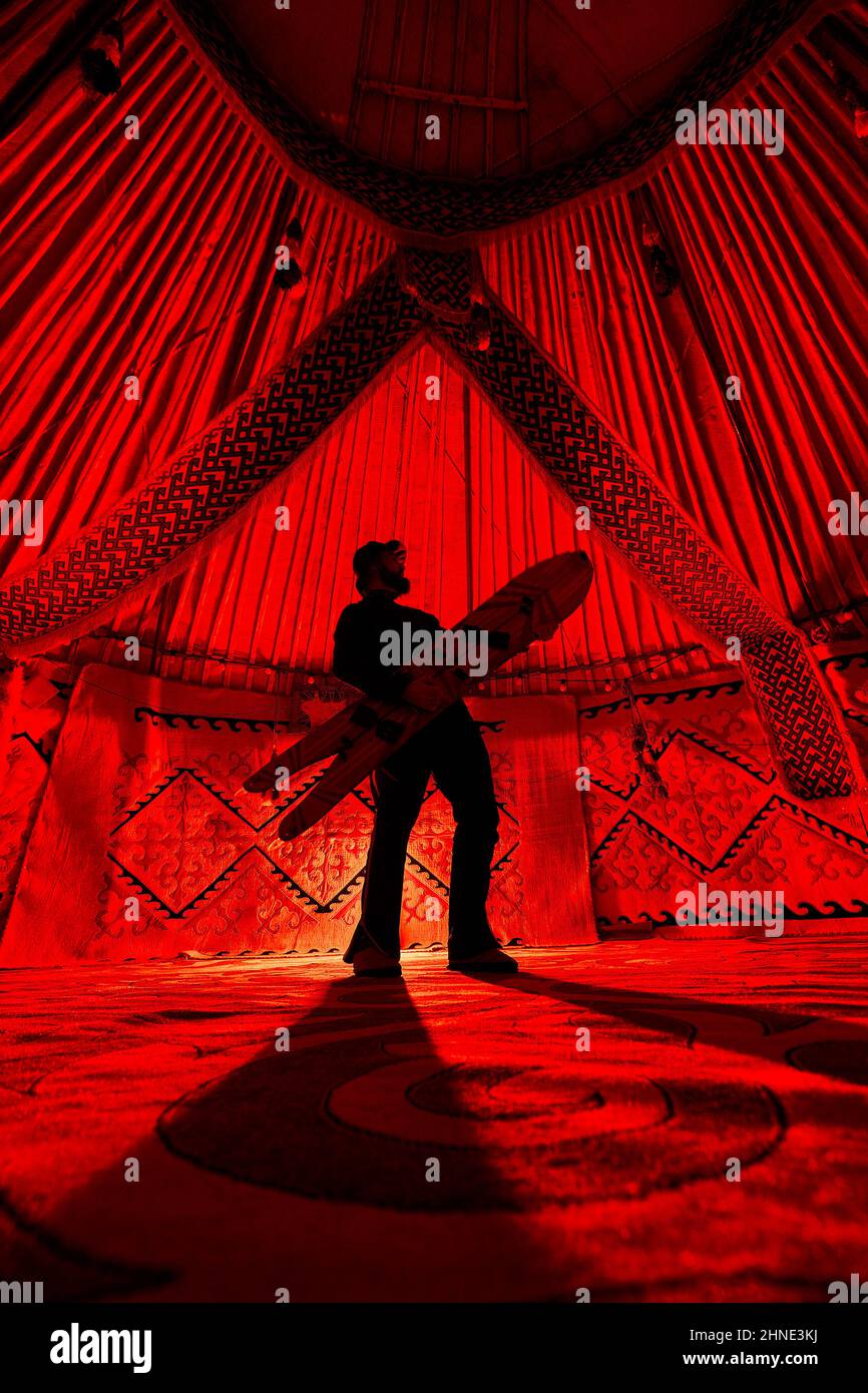 Silhouette von man mit gespaltener Schneebordwand wie Gitarre im Yurt Nomadenhaus gegen Teppich mit ethnischen Mustern, die von rotem Licht im Inneren beleuchtet werden. Backco Stockfoto