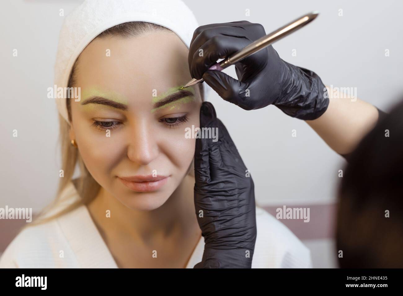 Die Hand eines Make-up Künstlers in schwarzen Handschuhen trägt eine gelbe Augenbrauenpaste auf die Augenbraue auf. Professionelle stilvolle dauerhafte Make-up, Augenbrauen färben Stockfoto