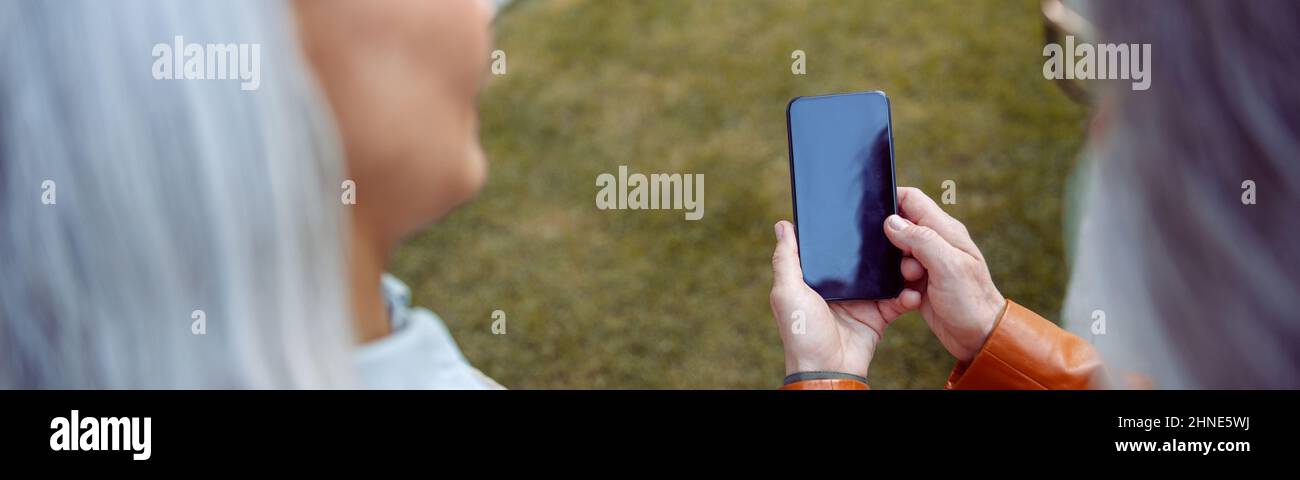 Reife Frau zeigt Handy mit leerem Bildschirm einem silberhaarigen Freund auf dem Rasen Stockfoto