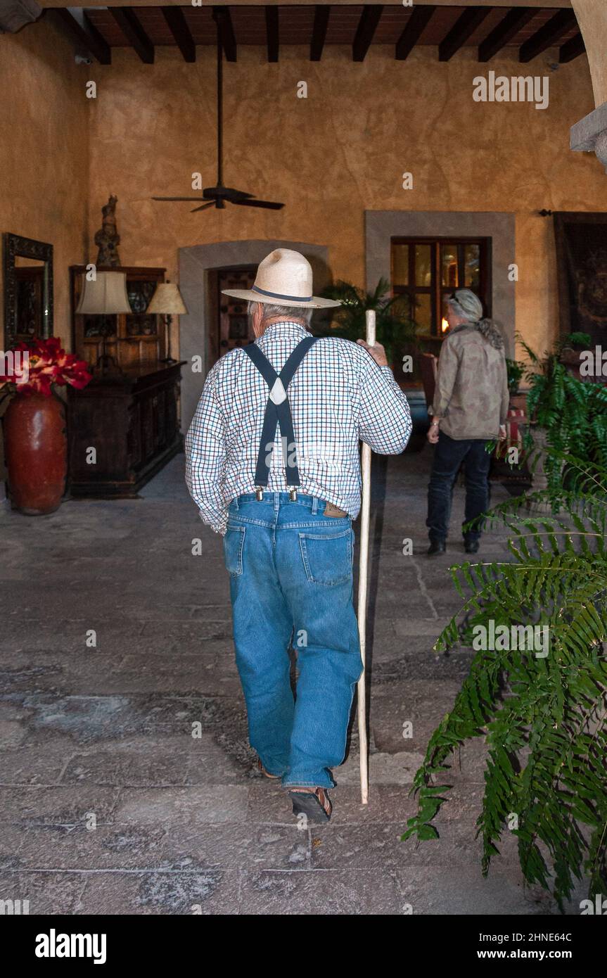 Ein einfacher, volkstümlicher älterer Mann in blauen Jeans, Hosenträgern und Strohhut schlendert mit einem Stock durch eine elegante Hotellobby im spanischen Stil in Mexiko. Stockfoto