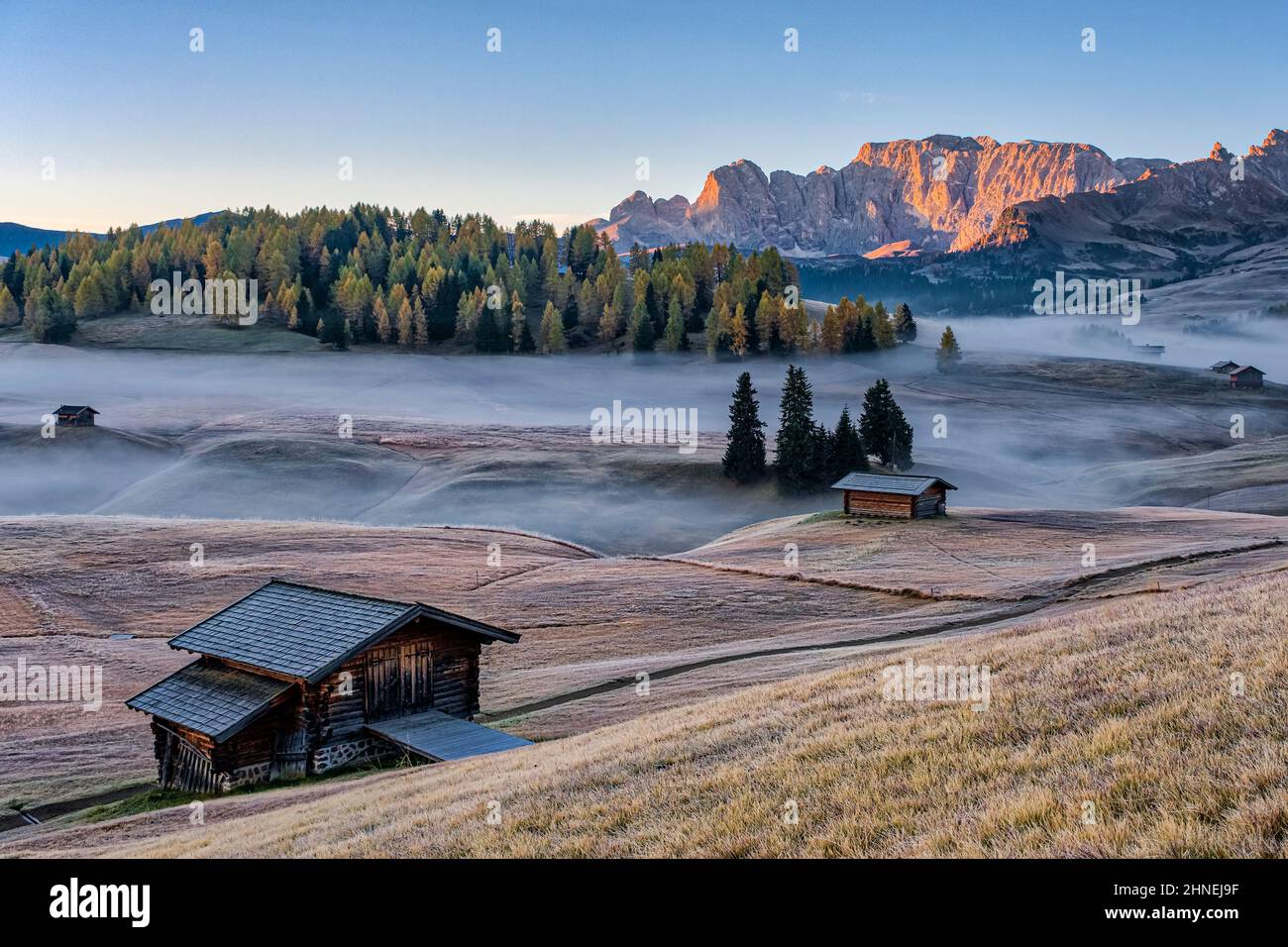 Nebel über den Weiden der Seiser Alm mit Holzhütten, die Roterdspitze in der Ferne, im Herbst bei Sonnenaufgang. Stockfoto