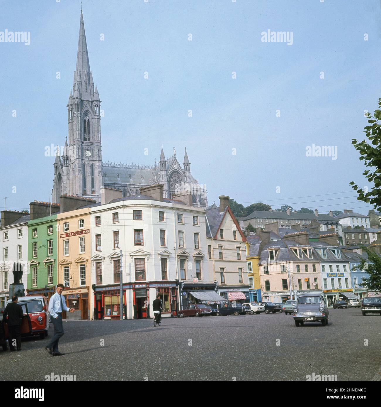 1960s, historischer Blick aus dieser Ära des Casement Square in Cobh, Co Cork, Irland, mit dem hohen Turm der St. Colman's Cathedral dahinter und Autos und Gebäuden, darunter Pubs, Ryans's und die Seagull Bar. Die Kirche von St. Colman's ist eines der höchsten Gebäude in Irland. Stockfoto