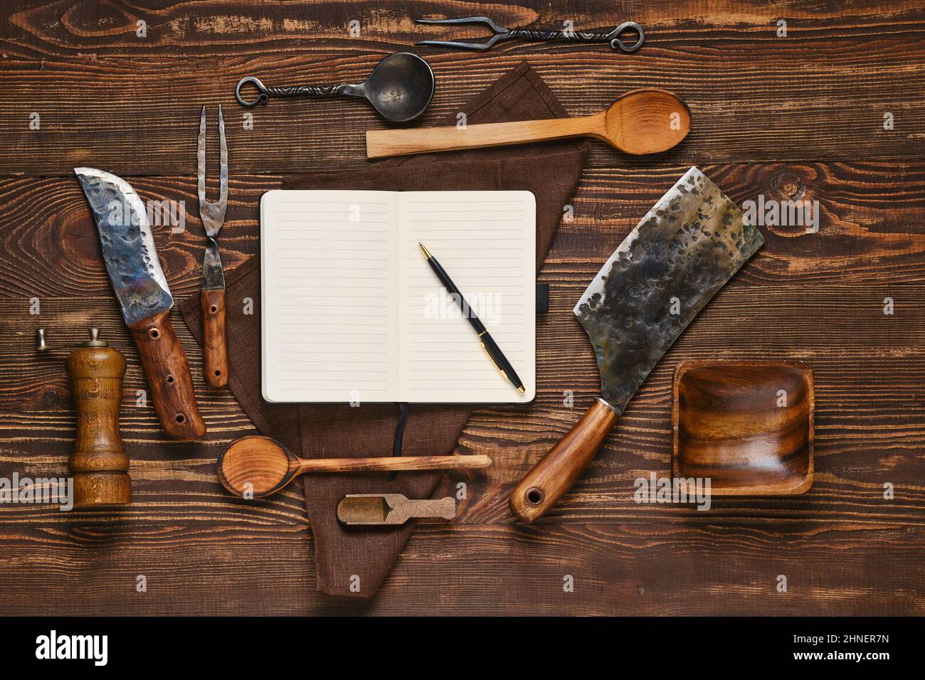 Draufsicht auf Vintage-Steakgabel, Messer und Beil auf Holztisch neben Notizbuch. Grillgeräte und offenen Notizblock, Food Blogging und Menü conce Stockfoto