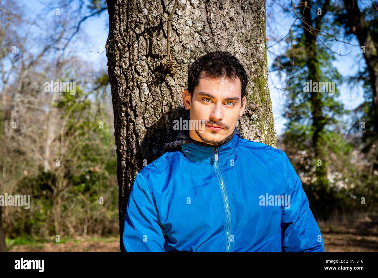 Porträt eines schönen jungen Mannes mit blauen Augen. Der Mann trainiert im Wald und lehnt sich an eine Eiche. Stockfoto