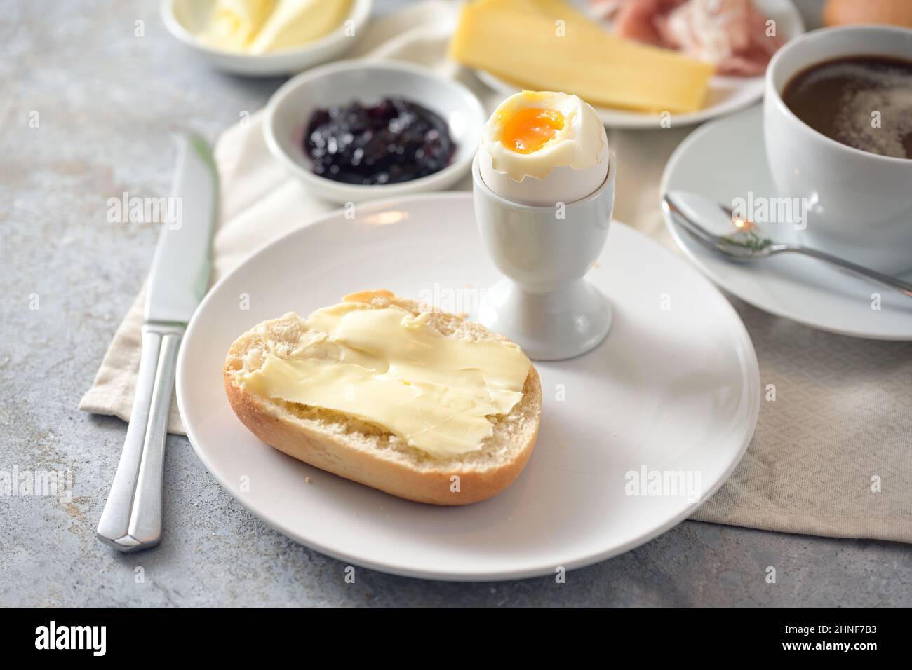 Brötchen mit Butter, gekochtem Ei, Marmelade und Kaffee auf einem Frühstückstisch, ausgewählter Fokus, enge Schärfentiefe Stockfoto