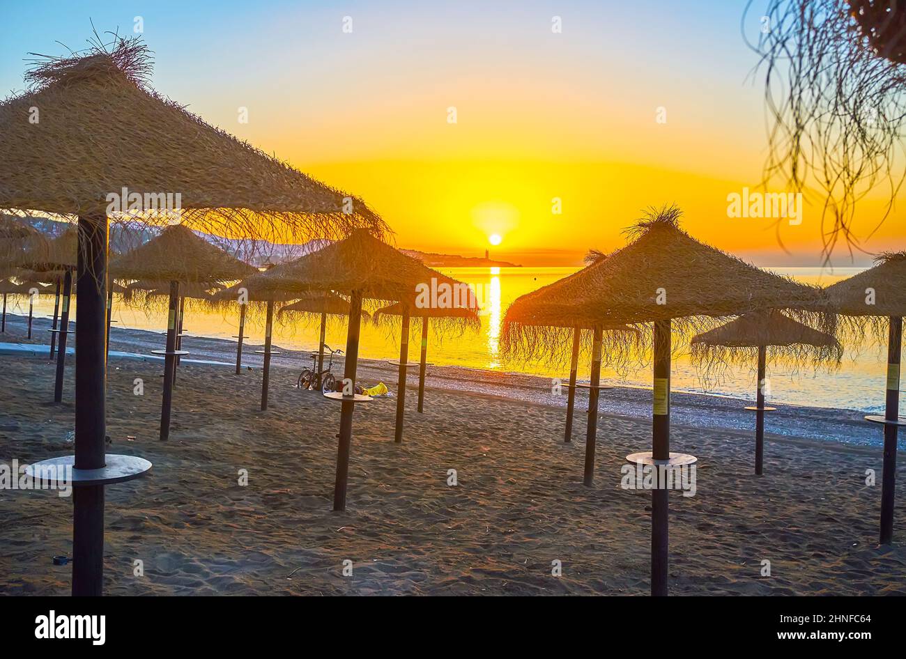 Der Sonnenaufgangsspaziergang am Strand von Malagueta mit Blick auf die zahlreichen Strohsonnenschirme, die Küste, den Sonnenaufgang und den Sonnenweg am Mittelmeer, Malaga, Stockfoto