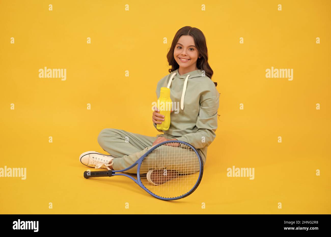 Glückliches Kind sitzt in Sportbekleidung mit Tennis-Schläger Wasserflasche auf gelbem Hintergrund, Wasserhaushalt Stockfoto