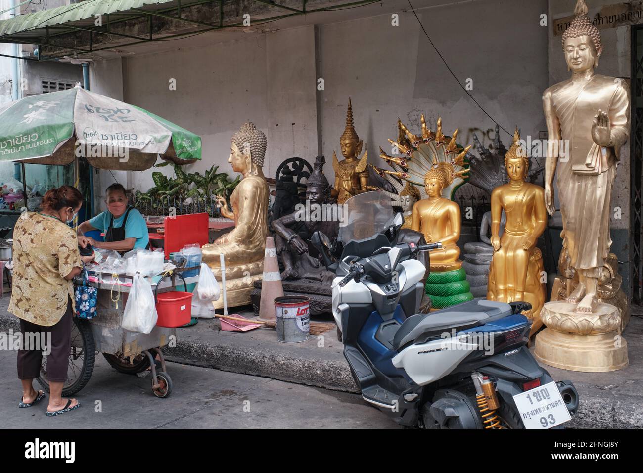 Ein Street-Food-Händler, der von ihrem Wagen aus vor verschiedenen Buddha-Statuen eines nahe gelegenen Ladens für religiöse Objekte operiert; Bangkok, Thailand Stockfoto
