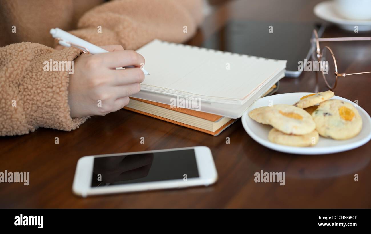 Eine Studentin der Millennials, die sich Notizen auf ihrem spiralförmigen Notizblock machte, während sie im Café Kekse aß. Stockfoto