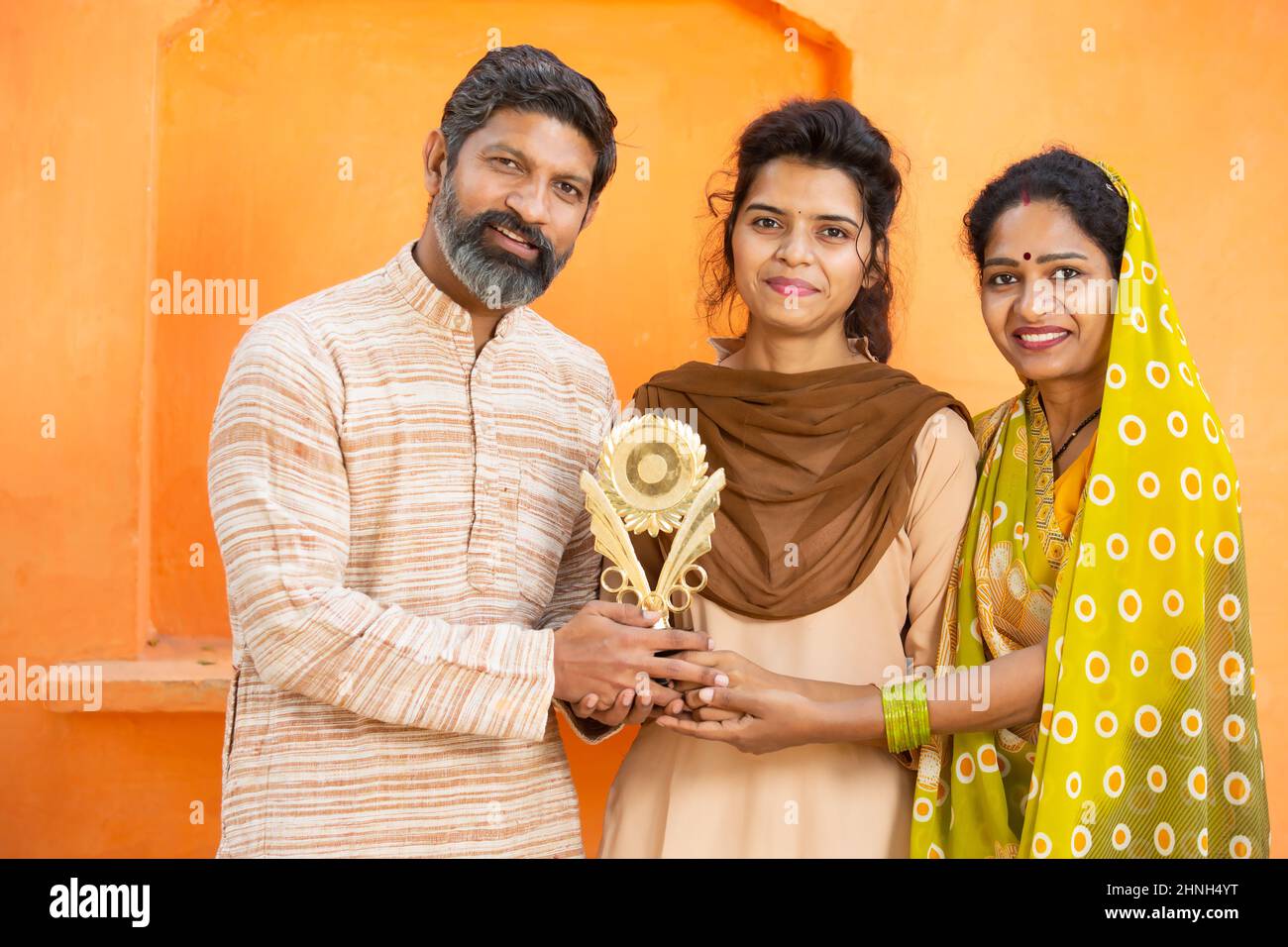 Stolze traditionelle indische Eltern mit einer jungen Tochter, die den Siegerpreis hält und den Sieg feiert. Glücklich junge Schulmädchen zeigt Trophäe stehen mit h Stockfoto