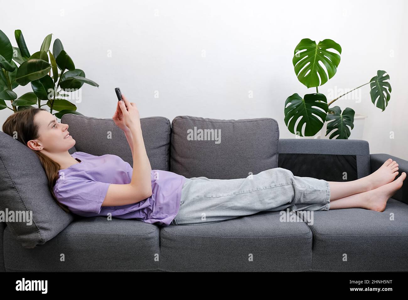 Seitenansicht einer ruhigen jungen Frau, die auf dem Sofa im Wohnzimmer liegt und ihr Smartphone in der Hand hält, schönes Mädchen, das sich chattet, soziale Netzwerke nutzt, Waren auswählt Stockfoto