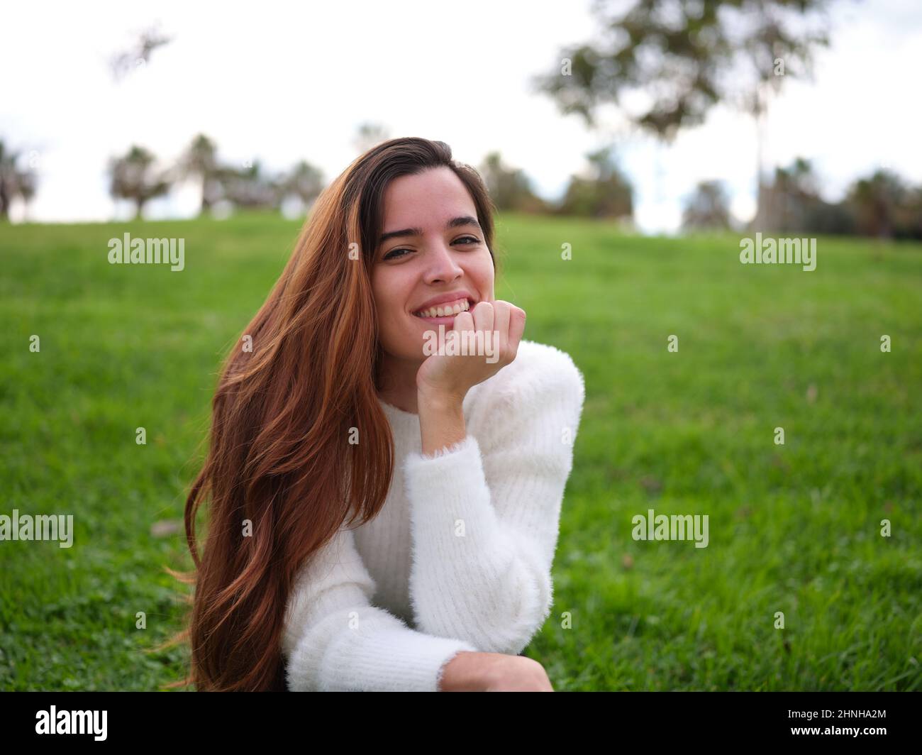 Eine junge Frau im Park lächelt fröhlich mit der Hand auf ihrem Kinn an der Kamera. Stockfoto