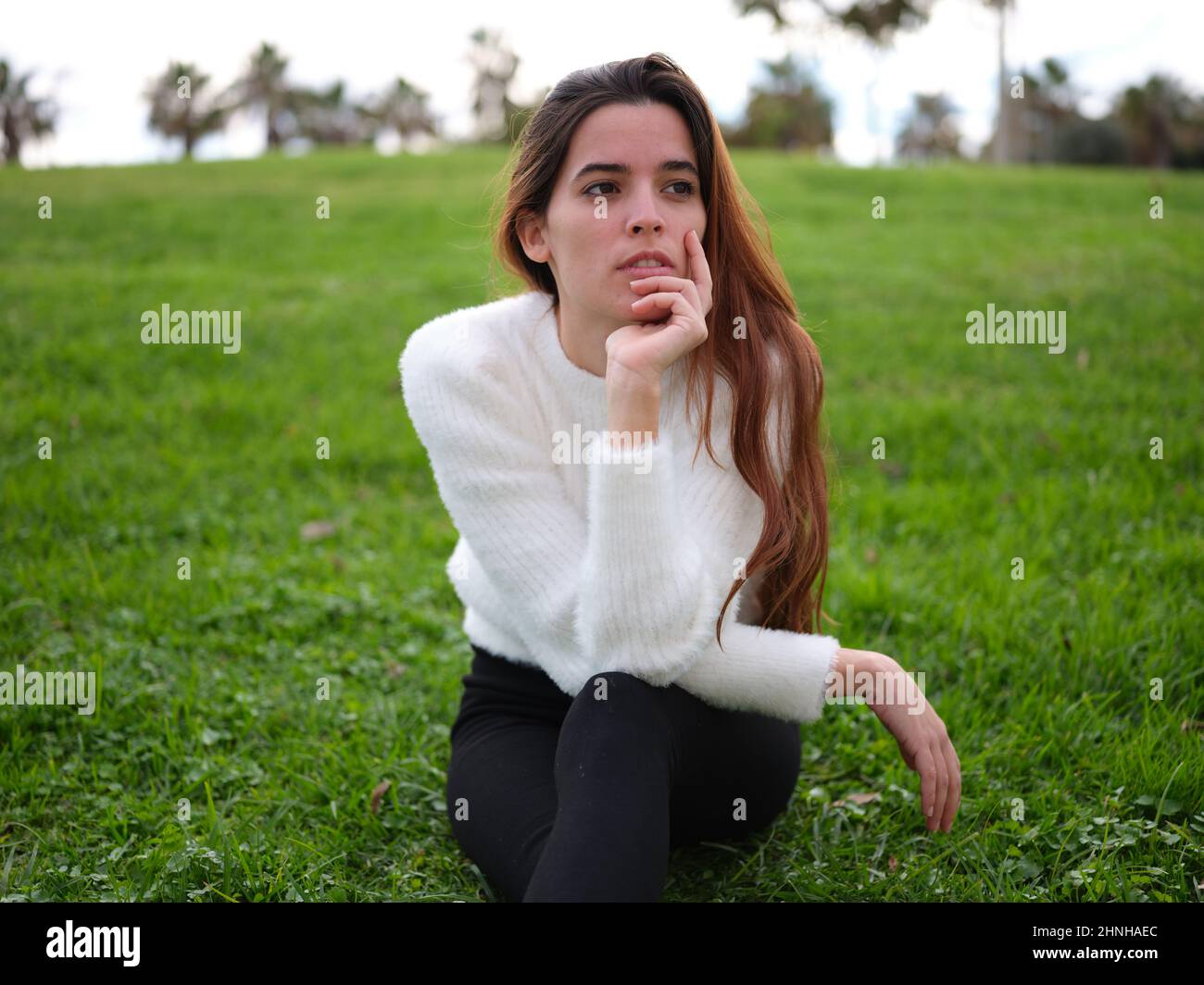 Eine nachdenkliche junge Frau, die im Park auf dem Gras sitzt und mit der Hand ins Gesicht blickt Stockfoto