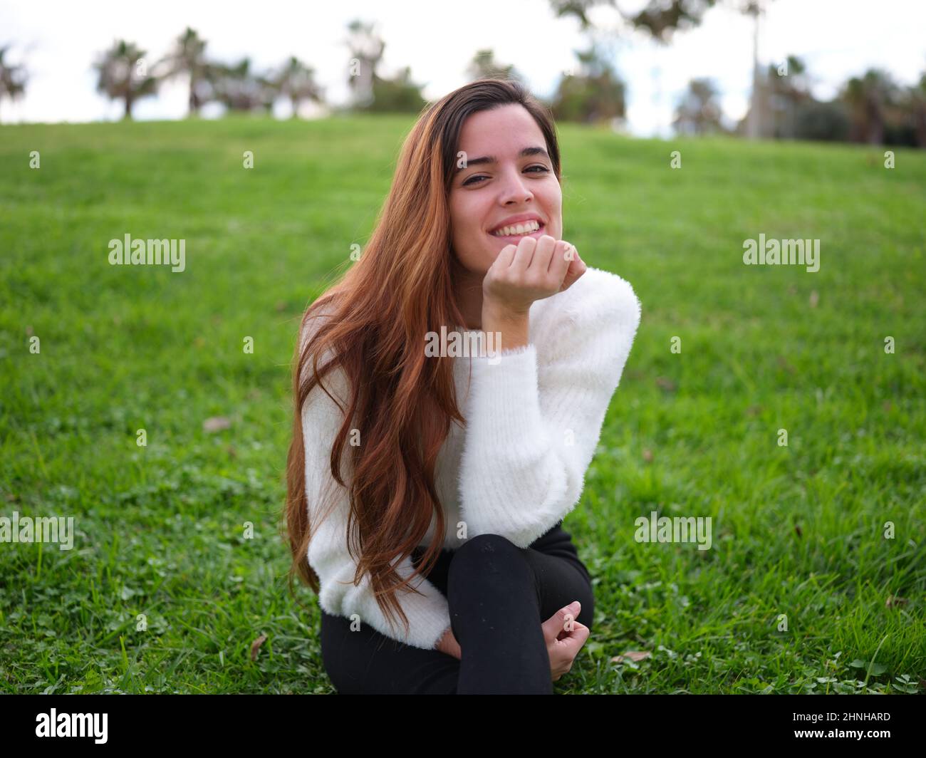 Eine verliebte junge Frau, die im Park auf dem Gras sitzt und mit der Hand auf dem Kinn die Kamera anlächelt. Stockfoto
