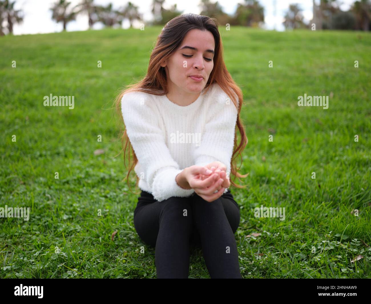 Eine schüchterne junge Frau, die mit geschlossenen Augen auf dem Gras im Park sitzt und ihre Hände an ihren Beinen zusammenhält. Stockfoto
