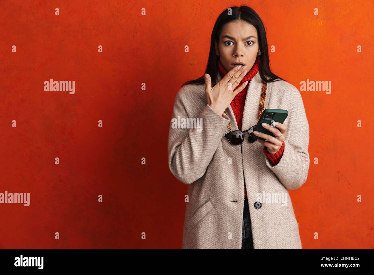 Schockieren sie die hispanische Frau, die ihren Mund bedeckt, während Sie ein über einer orangen Wand isoliertes Mobiltelefon benutzen Stockfoto