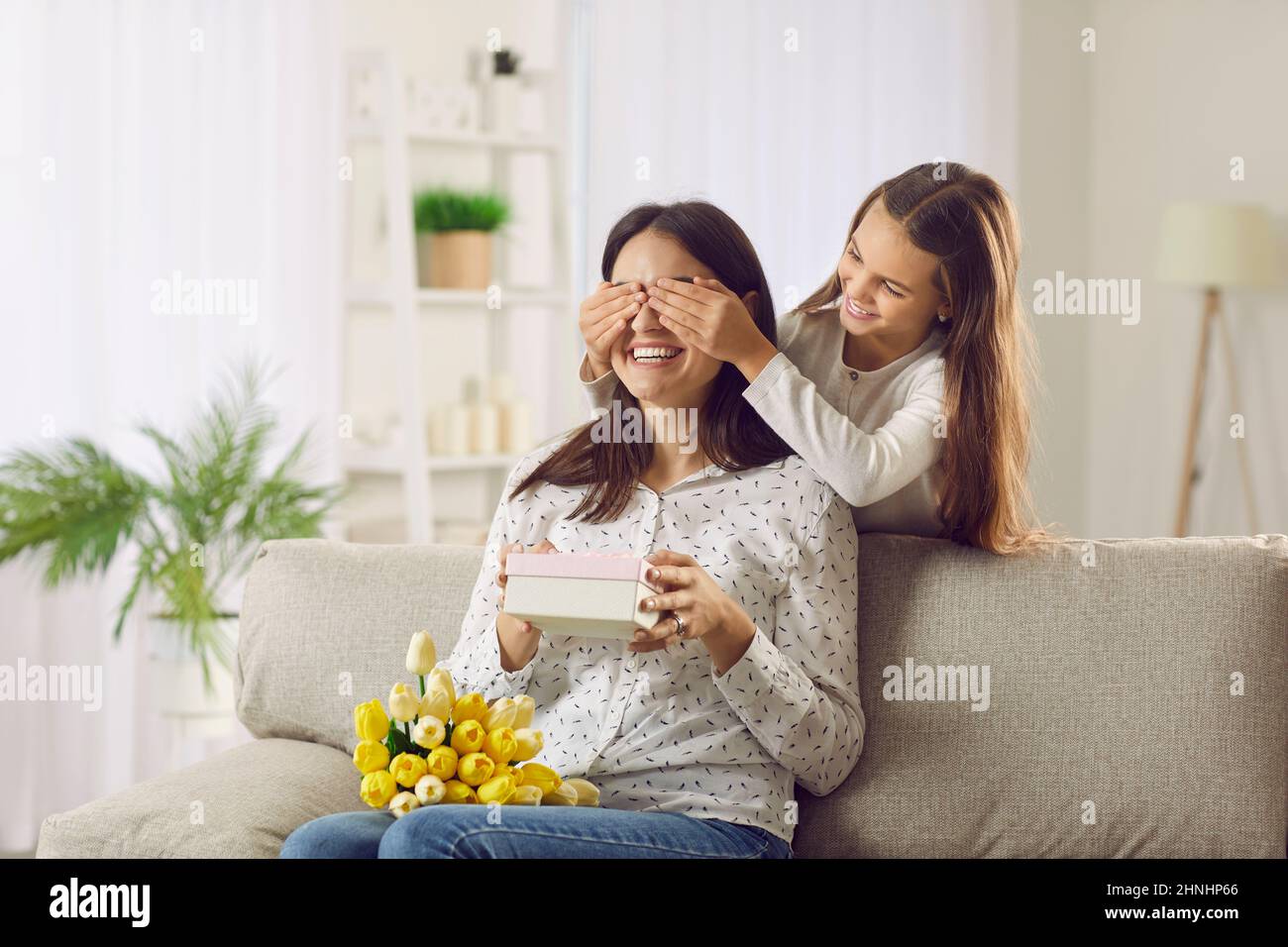 Die kleine Tochter bedeckt die Augen ihrer Mutter, als sie ihr am Muttertag ein Geschenk macht Stockfoto