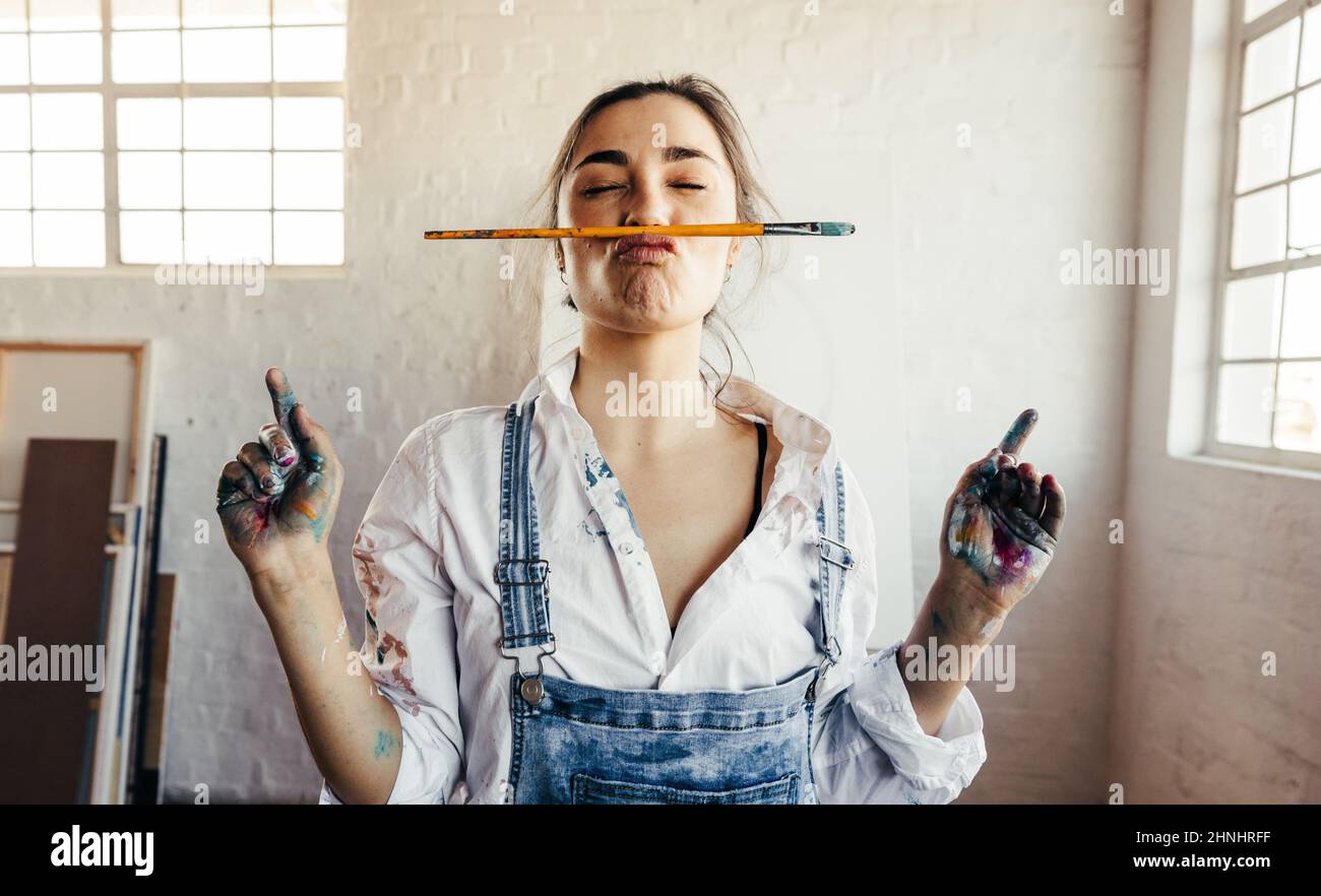 Verspielte Malerin, die mit einem Pinsel ein lustiges Gesicht macht. Die junge Künstlerin steht in ihrem Atelier mit ihrer leeren Leinwand im Hintergrund. Stockfoto