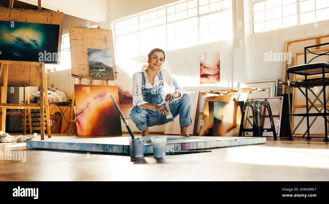 Kreative Malerin, die in einem Kunststudio nahe an ihre Malerei hockt. Künstlerische junge Frau, die an einem neuen Gemälde auf dem Boden arbeitet. Fröhlicher junger Künstler Stockfoto
