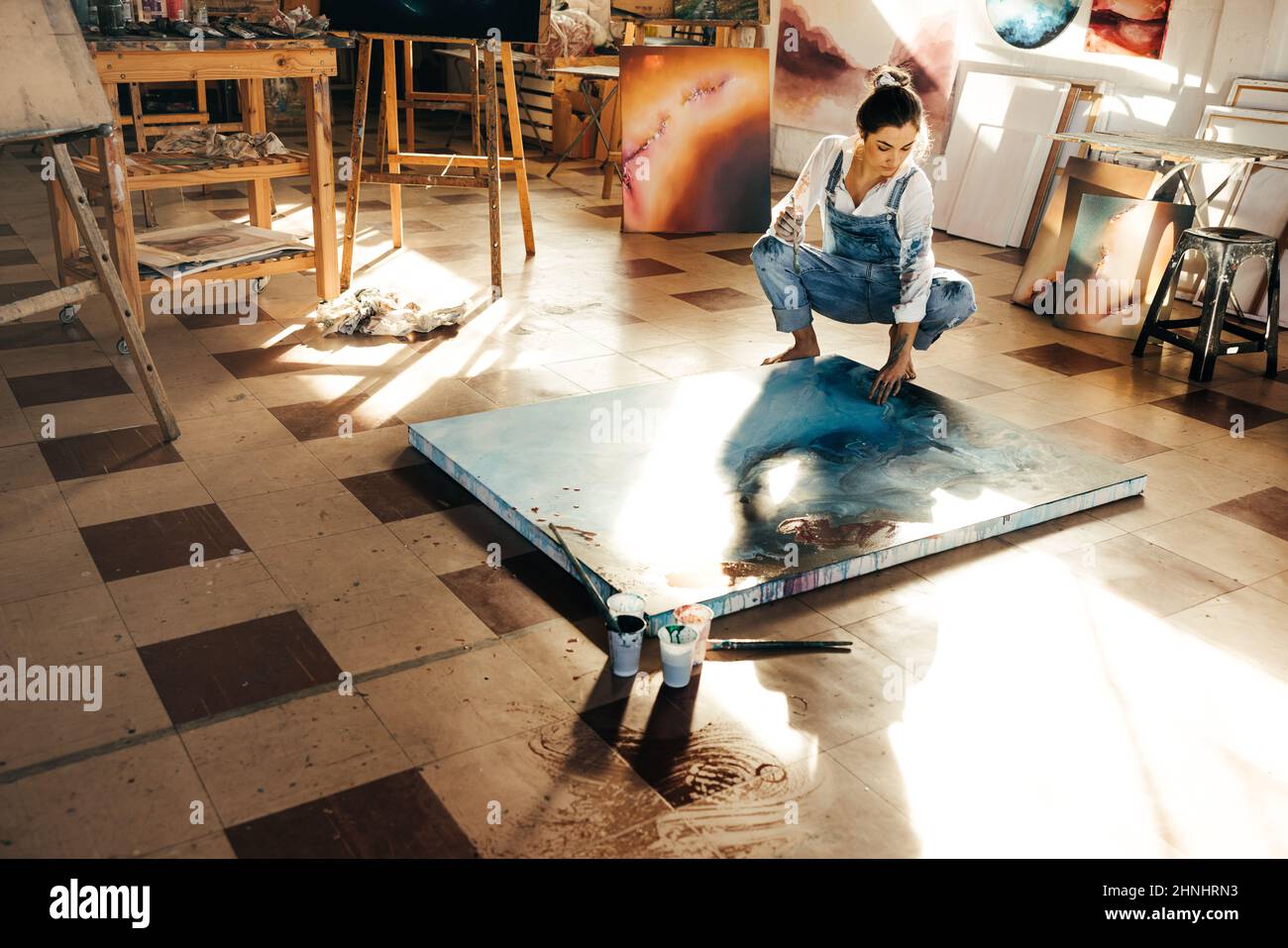 Malerin, die ein blaues Gemälde auf einer Leinwand malt. Kreative junge Frau berührt ihr Gemälde, während sie in ihrem Kunststudio auf dem Boden hockt. Künstlerisch Stockfoto