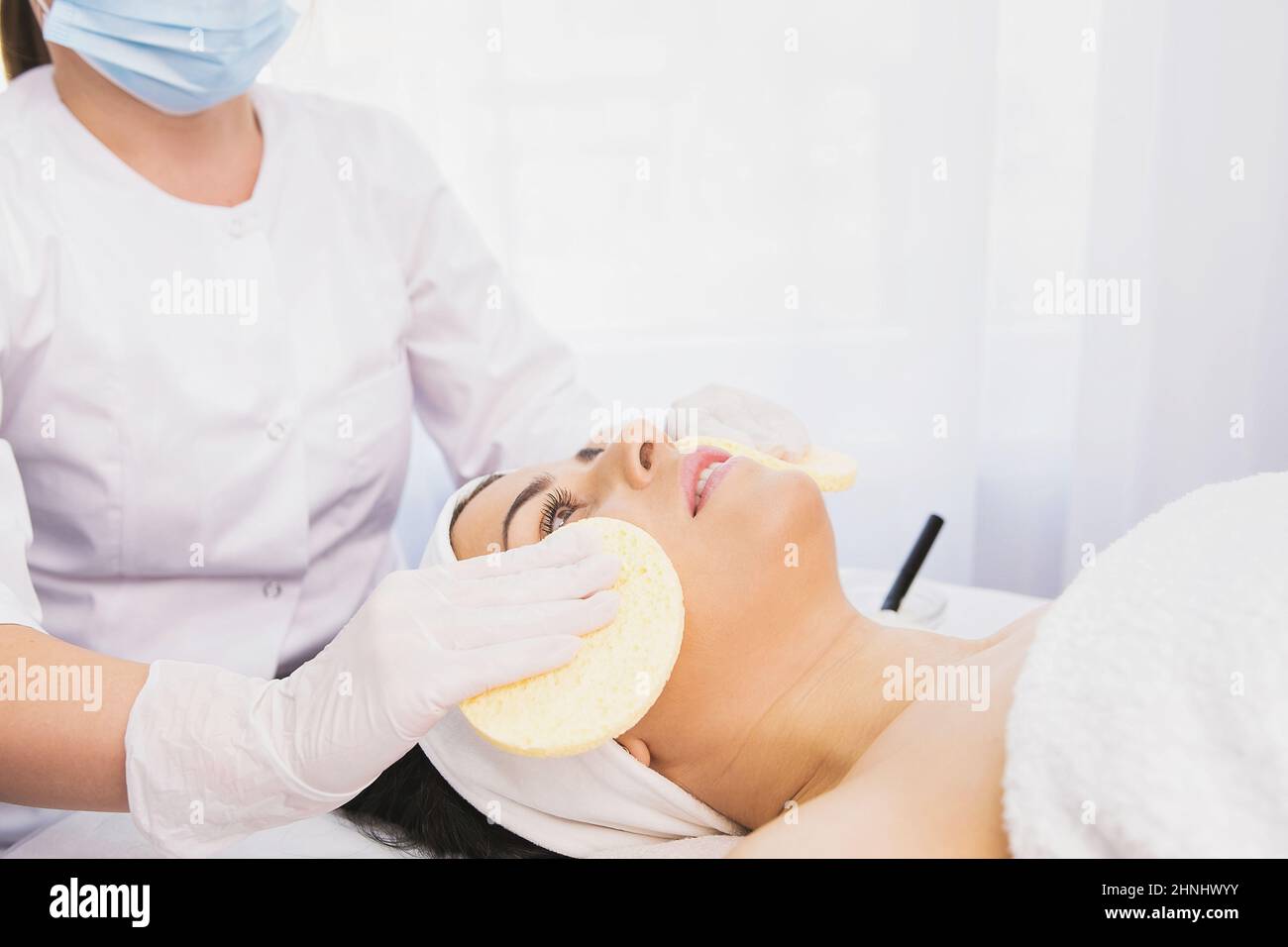 Kosmetikerin in medizinischen Einweg-Maske und Handschuhe reinigt die Haut der schönen Frau mit einem Schwamm, Spa-Behandlung Hautpflege Gesicht. Reinigung und Tonisierung Stockfoto