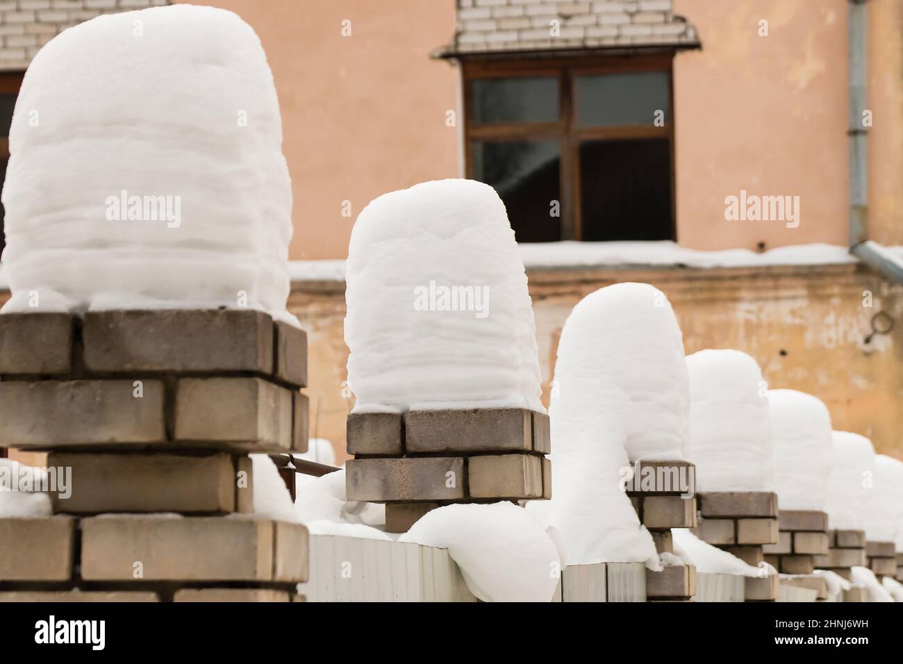 Hohe, wunderschöne Säulen aus weißem Schnee auf Ziegelsteinsäulen. Eine gerade Reihe kleiner Schneeverwehungen vor dem Hintergrund einer schäbigen Wand. Urbane Winterlandschaft. Wolkiger Wintertag, weiches Licht. Stockfoto