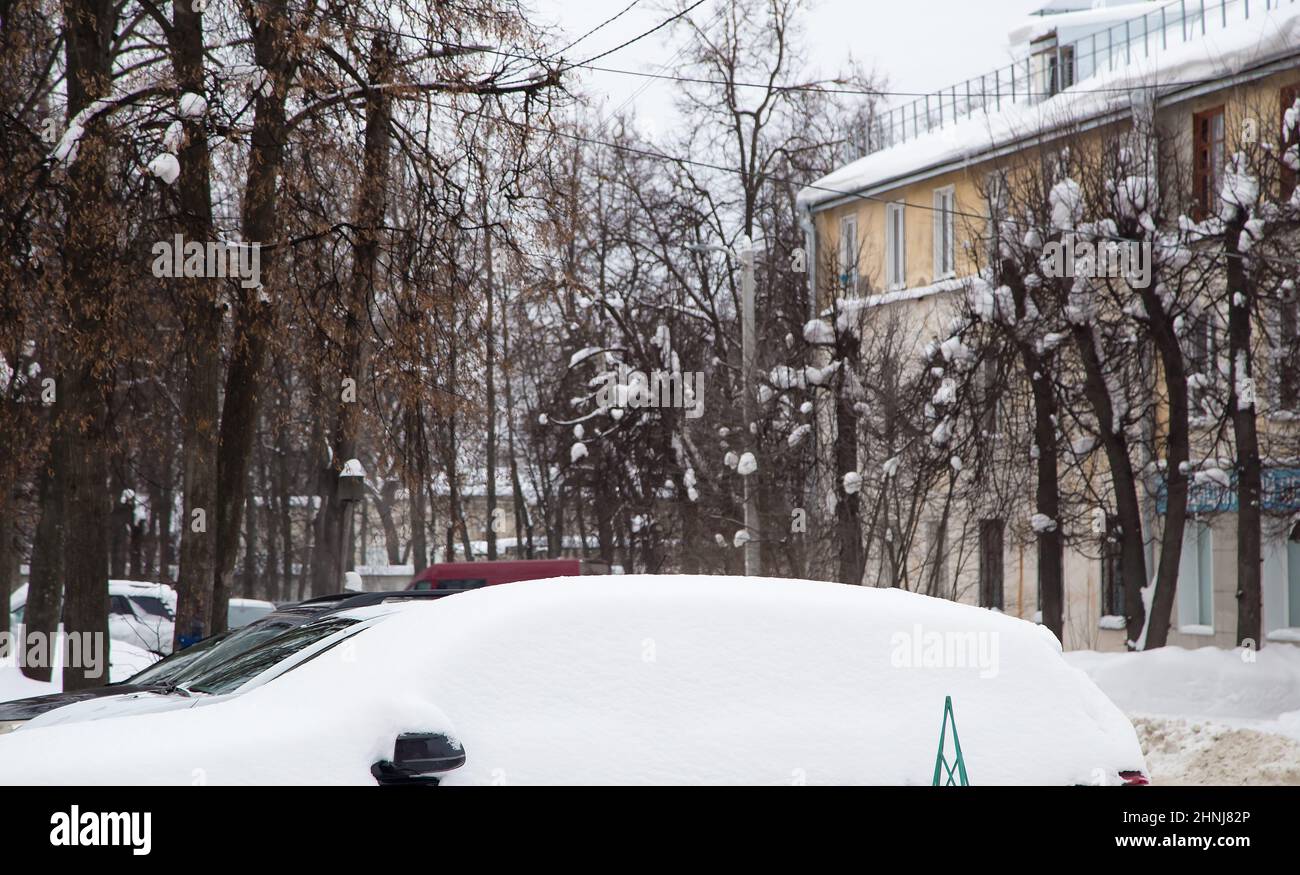 Eine lockere Schneeverwehung von Autos an der Straße vor dem Hintergrund einer Stadtstraße. Auf der Straße liegt schmutziger Schnee in hohen Haufen. Urbane Winterlandschaft. Wolkiger Wintertag, weiches Licht. Stockfoto