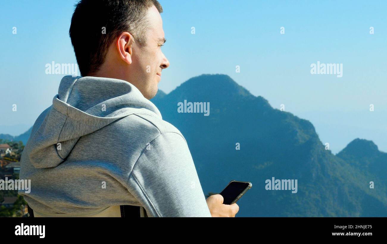 Reisemann mit Mobiltelefon im Hoodie sitzend und genießen die Aussicht auf den Berg Stockfoto
