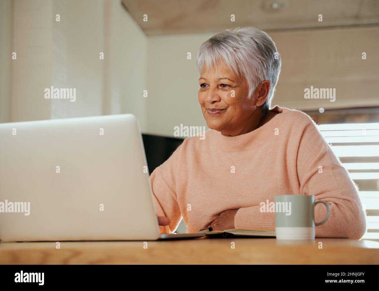 Ältere, multiethnische Frauen, die am heimischen Tresen mit einem Laptop sitzen und lächeln. Stockfoto