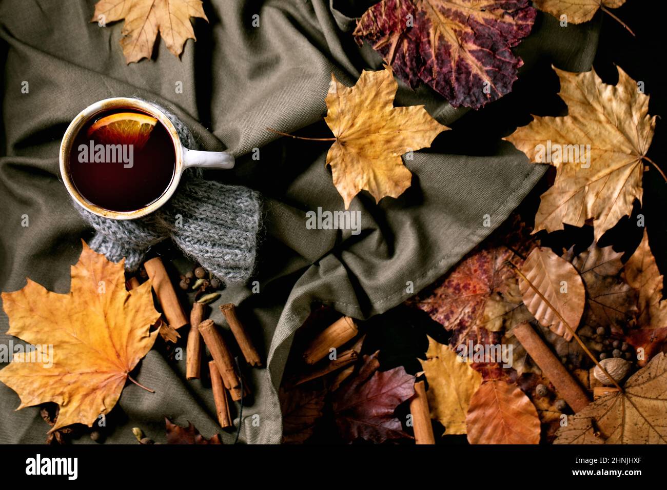 Heißer Glühwein im Keramikbecher im Schal mit Gewürzen, Orange und Herbstblättern über dunkler Leinentischdecke. Flach liegend. Gemütliche warme Getränke. Flach liegend Stockfoto