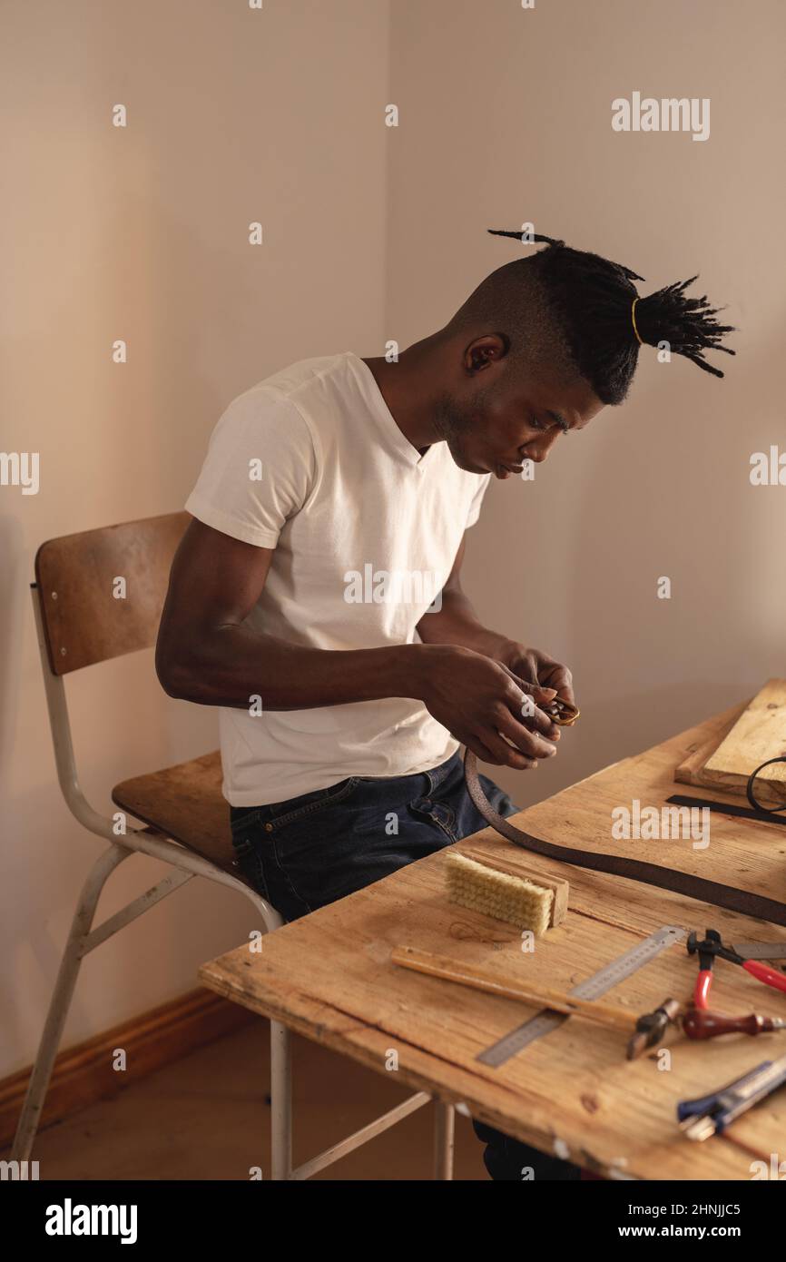 afroamerikanischer junger Handwerker, der während der Arbeit in der Werkstatt eine Schnalle am Ledergürtel befestigt Stockfoto