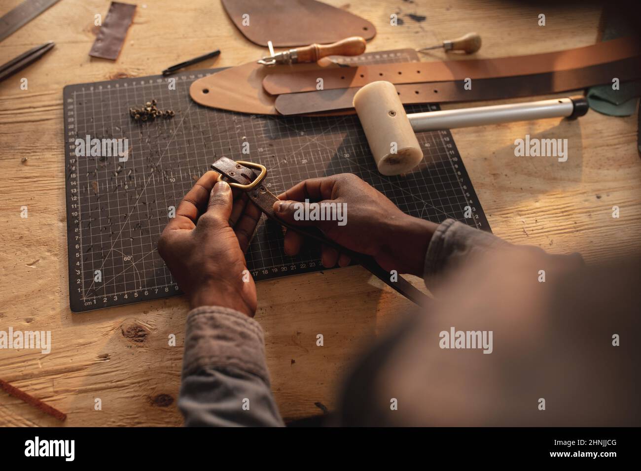 Beschnittene Hände eines jungen afroamerikanischen Handwerkers, der an der Werkbank eine Schnalle am Ledergürtel festhält Stockfoto