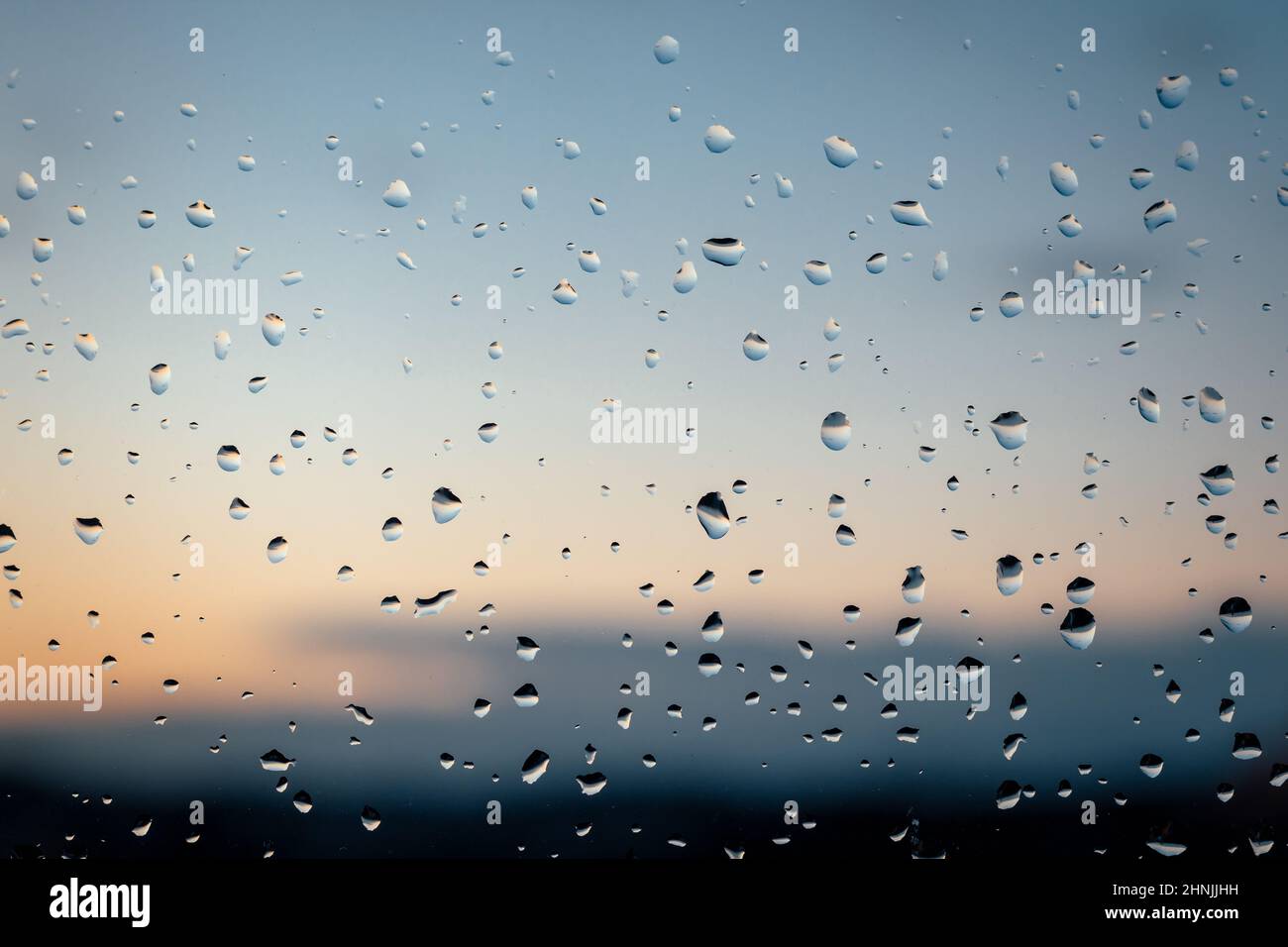 Regentropfen nach einem Sturm am Fenster bei Sonnenuntergang. Die Regentropfen sind fokussiert und der Hintergrund ist unscharf. Der Sonnenuntergang hat orange und rosa Farbtöne. Stockfoto
