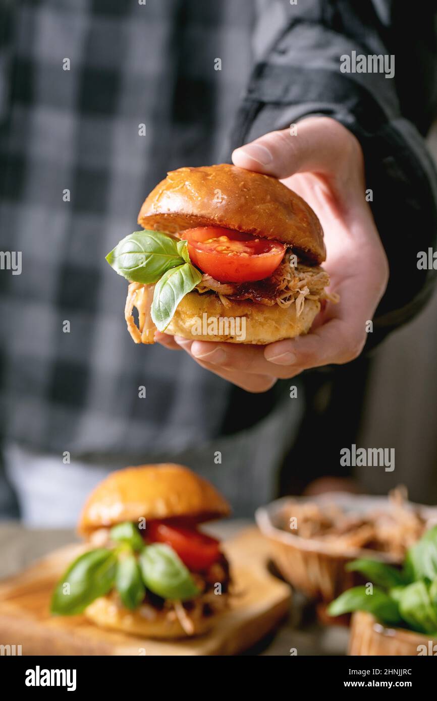 Der junge Chefkoch hält Mini-Hamburger mit Rindfleisch, Tomaten und Basilikum in der Hand über den Küchentisch mit anderen Hamburgern. Modern lecker schnell foo Stockfoto