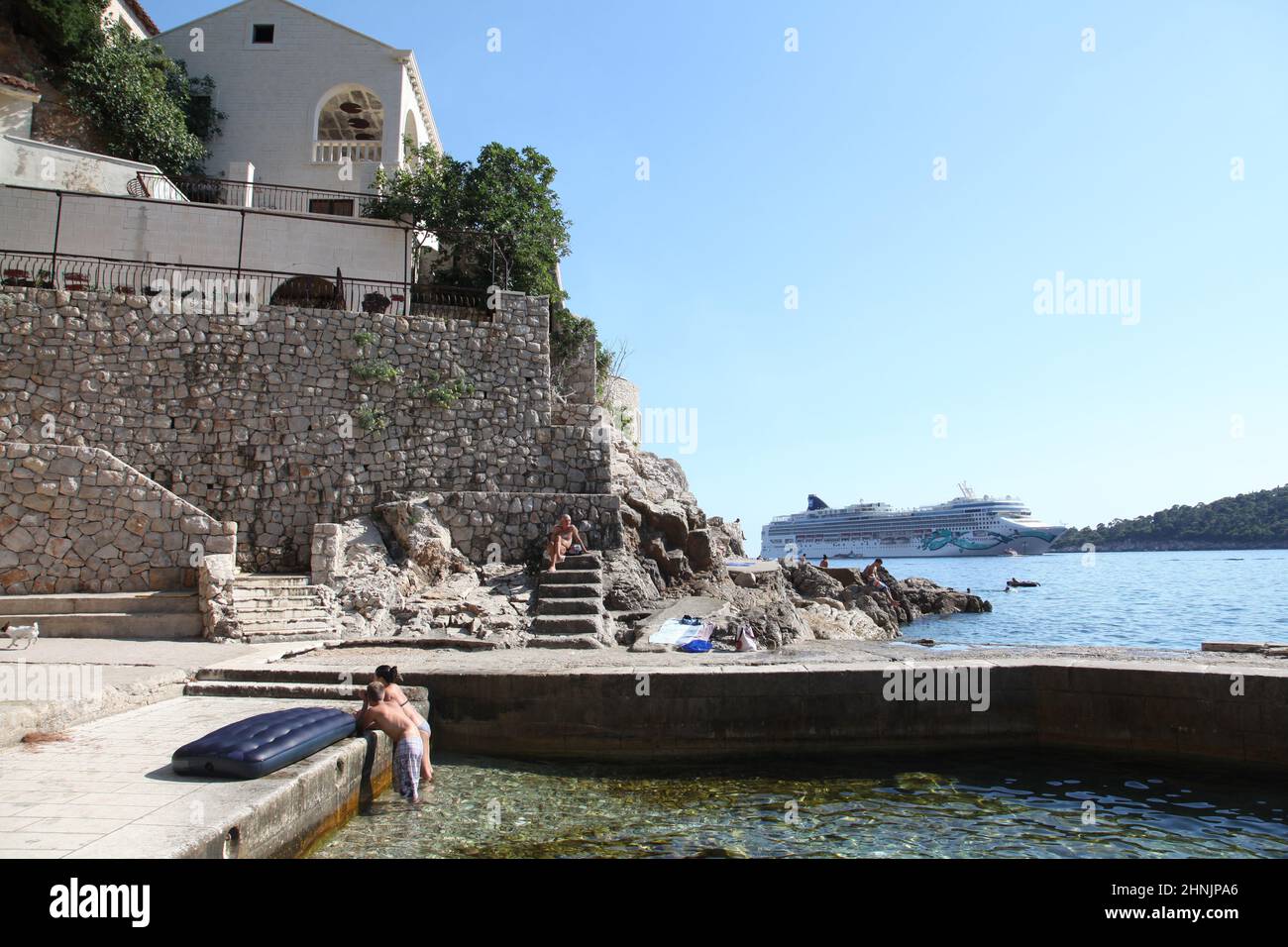 Das junge Paar genießt den Sommernachmittag am Strand von St. Jacob an der Adriaküste, während sich das Kreuzschiff Norwegian Jade dem Ankerplatz in der Lapad-Bucht von Dubrovnik nähert. Stockfoto