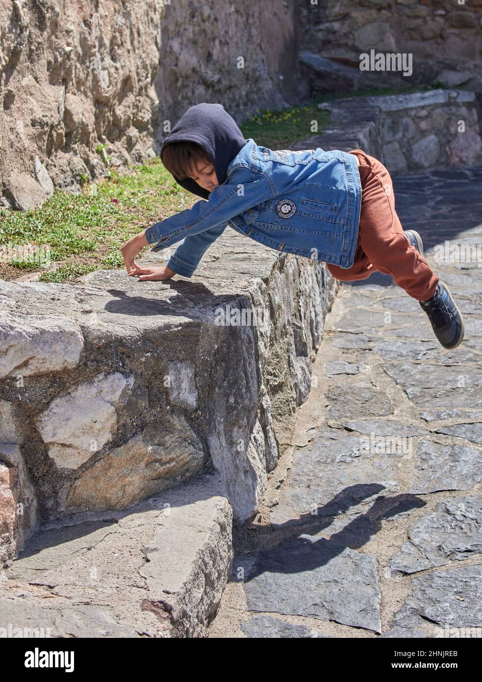 lateinisches Kind in lässiger Kleidung mit einer jean Kapuzenjacke, die Parkour übt. Kleiner Junge, der in einer niedrigen Mauer in der Altstadt Tricks macht Stockfoto
