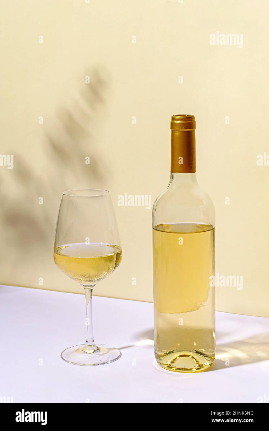 Unbeschriftete Weißweinflaschen mit halb leerem Weinglas, das auf einem weißen Tisch gegen eine neutrale Wand mit Werbeschilder steht Stockfoto