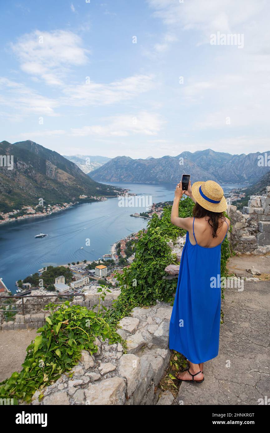 Ein Mädchen in blauem Kleid und Strohhut steht vor der Kulisse der wunderschönen Bucht von Kotor in Montenegro und fotografiert mit ihrem Handy. Stockfoto