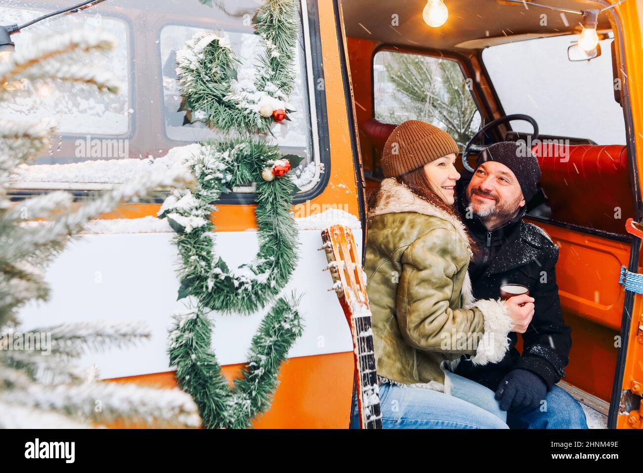 Glückliches, romantisches Paar mittleren Alters, verliebt, sitzt in einem Kleinbus, der mit Nadelbäumen geschmückt ist und heißen Tee trinkt. Mann und Frau in warmen Kleidern entspannen sich Stockfoto