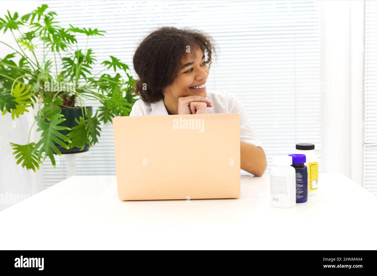 Junge glückliche afroamerikanische Ärztin, die am Arbeitsplatz sitzt, wegschaut und lächelt, während sie in einer modernen Klinik am Laptop arbeitet, erfreut ethnische Phys Stockfoto