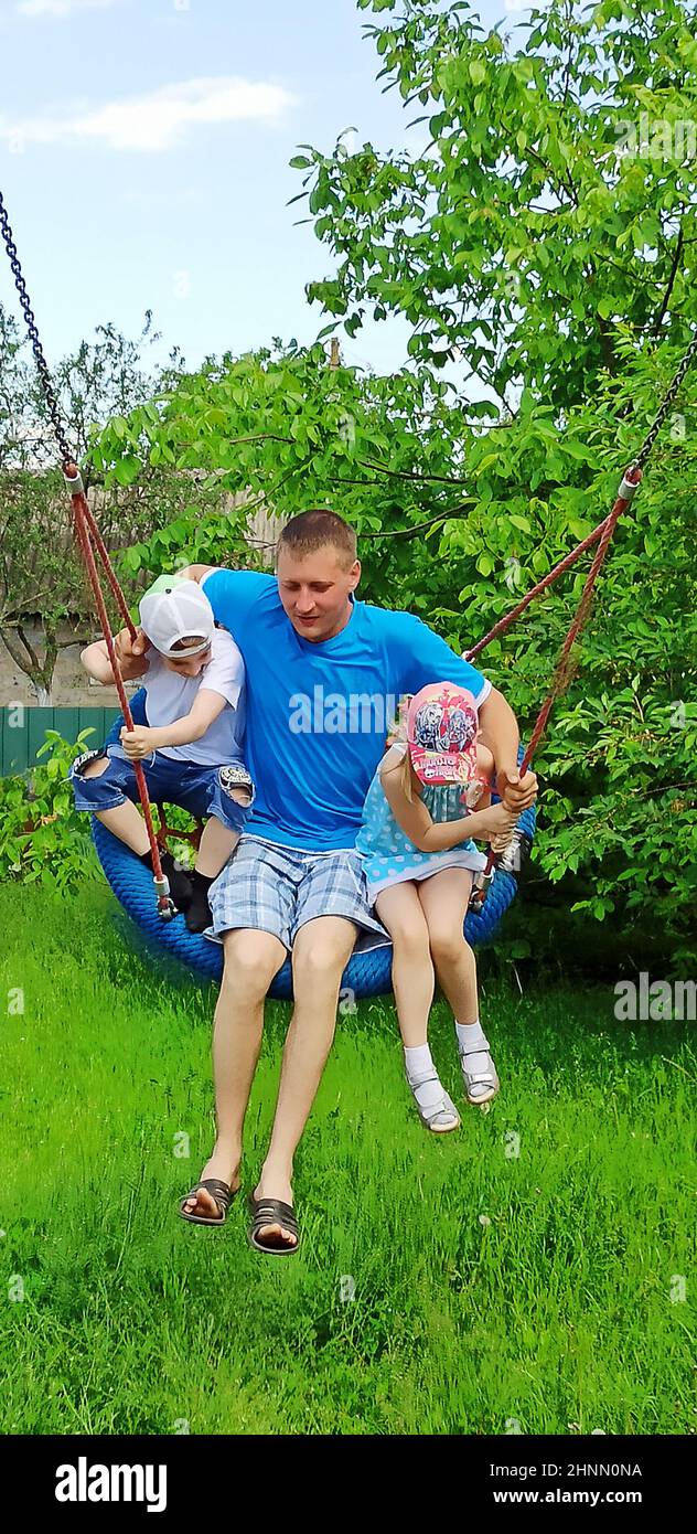 Glücklicher Vater mit Kindern, die Schaukel im Garten reiten. Familie genießt den Sommer Stockfoto