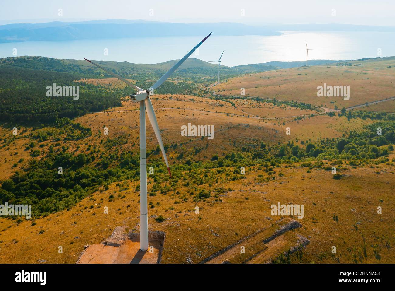 Windmühlen erzeugen grüne Energie an einer elektrischen Station auf einem mit Büschen bedeckten Hügel gegen Wälder in der Nähe des Adriatischen Meeres bei sonnigem Blick von der Drohne aus Stockfoto