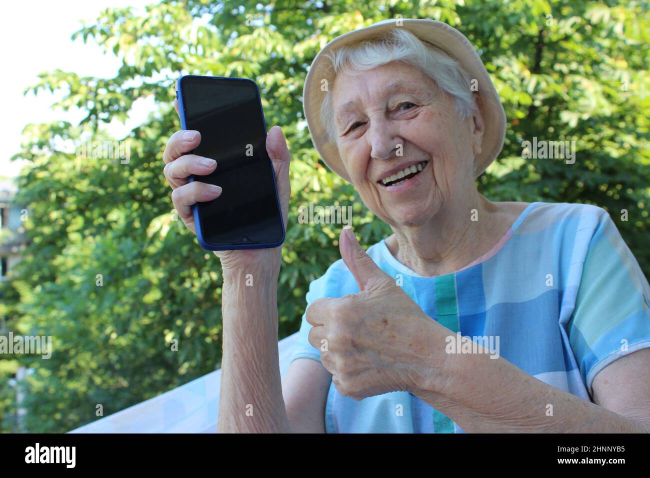 Fröhliche reife Dame zeigt ein Telefon isoliert auf grünen Baum Sommer Hintergrund Stockfoto