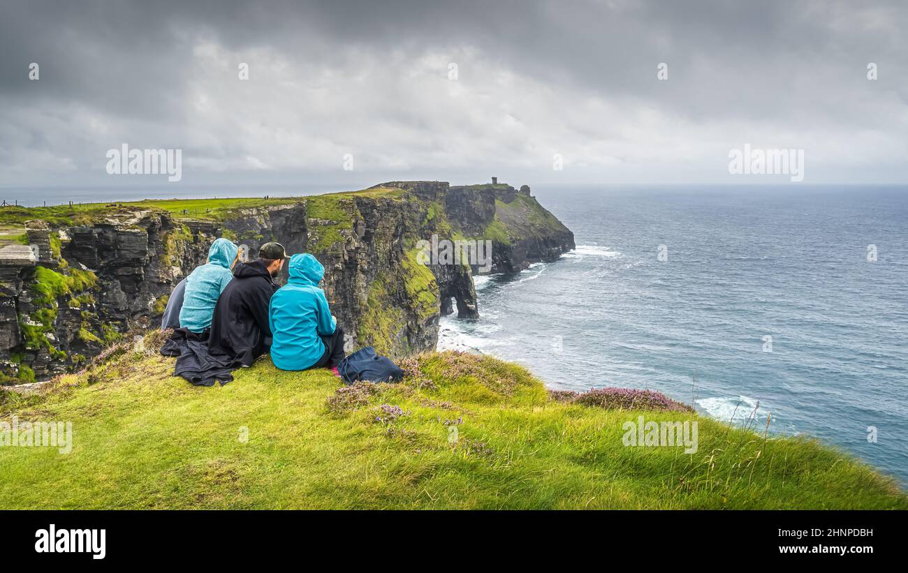 Eine Gruppe junger Menschen, die am Rand einer Klippe auf den berühmten Cliffs of Moher, Irland, sitzen Stockfoto