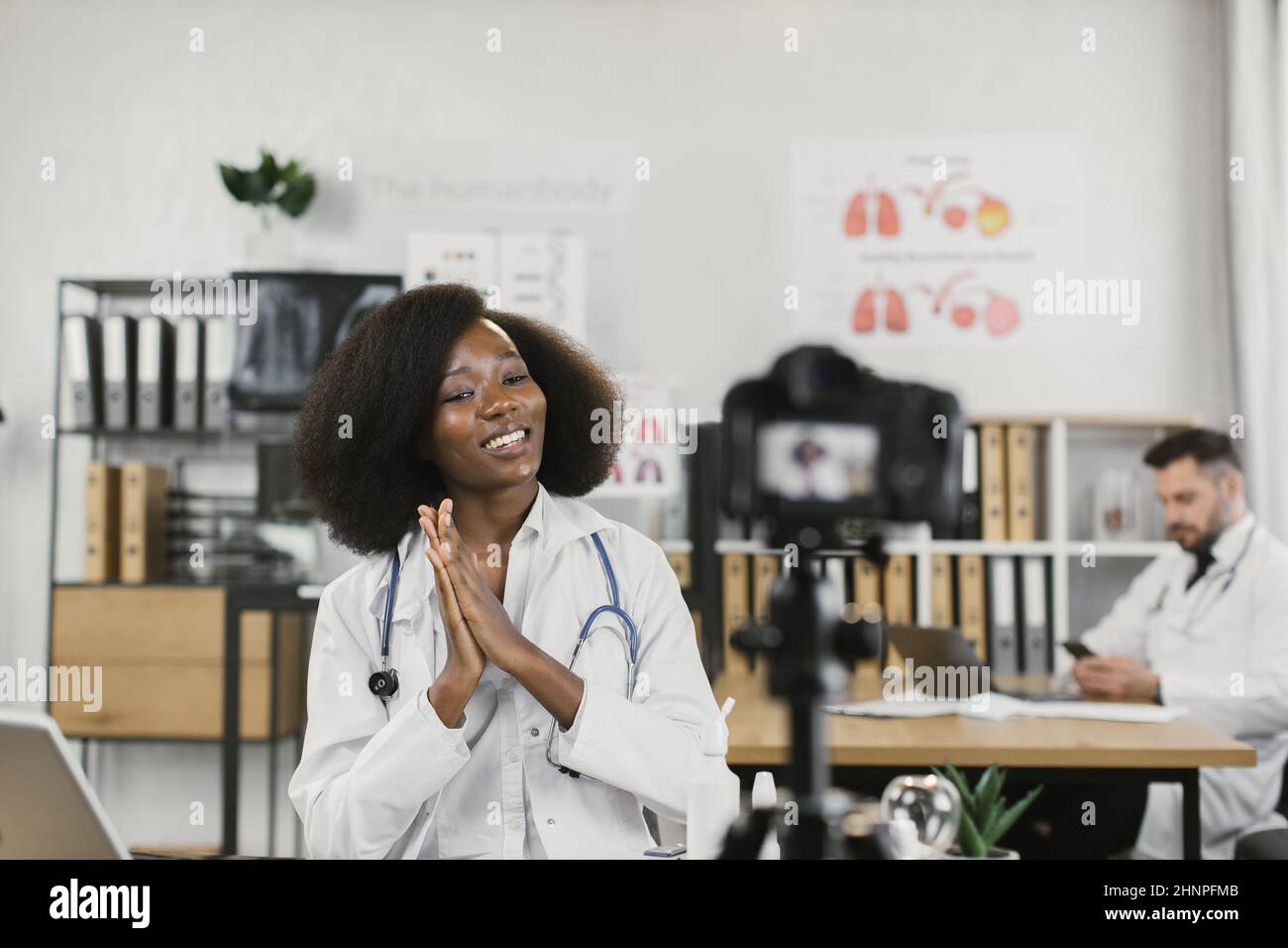 Afroamerikanische Frau im Laborkittel spricht und gestikiert, während sie einen Videoblog für ihren medizinischen Kanal aufzeichnet. Informativer Blog der kompetenten Ärztin. Stockfoto