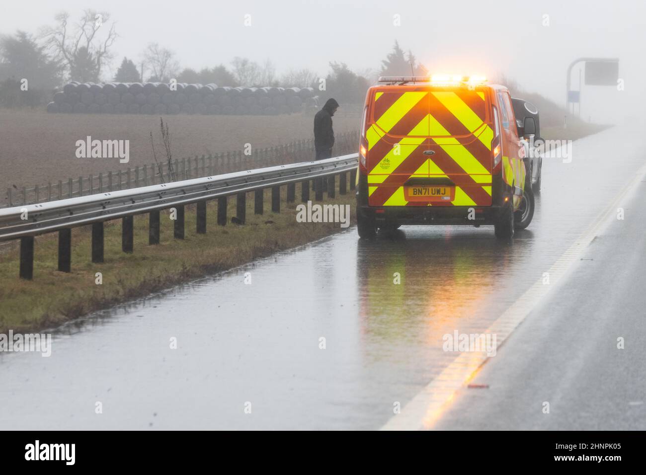 Mann, der hinter einer Autobahnabsperrung auf der britischen Autobahn steht, als RAC-Pannenfahrzeug und Mechaniker bei starkem Regen an seinem abgebrochenen Fahrzeug arbeiten - Großbritannien Stockfoto