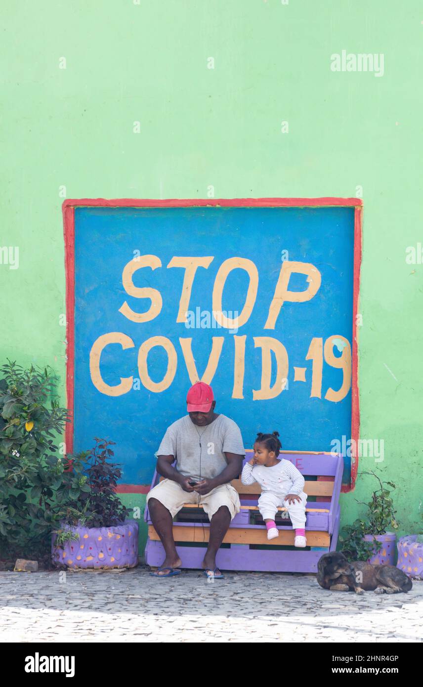 Vater und Kind sitzen unter dem Schild „Stop Covid-19“, Santa Maria, Sal, República de Cabo (Kap Verde) Stockfoto