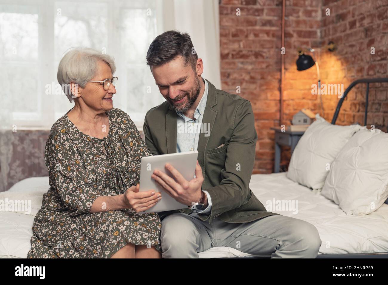 Ältere grauhaarige kaukasische Dame genießt ihre Zeit und schaut sich Fotos auf dem Tablet ihres Sohnes mittleren Alters an. Hochwertige Fotos Stockfoto