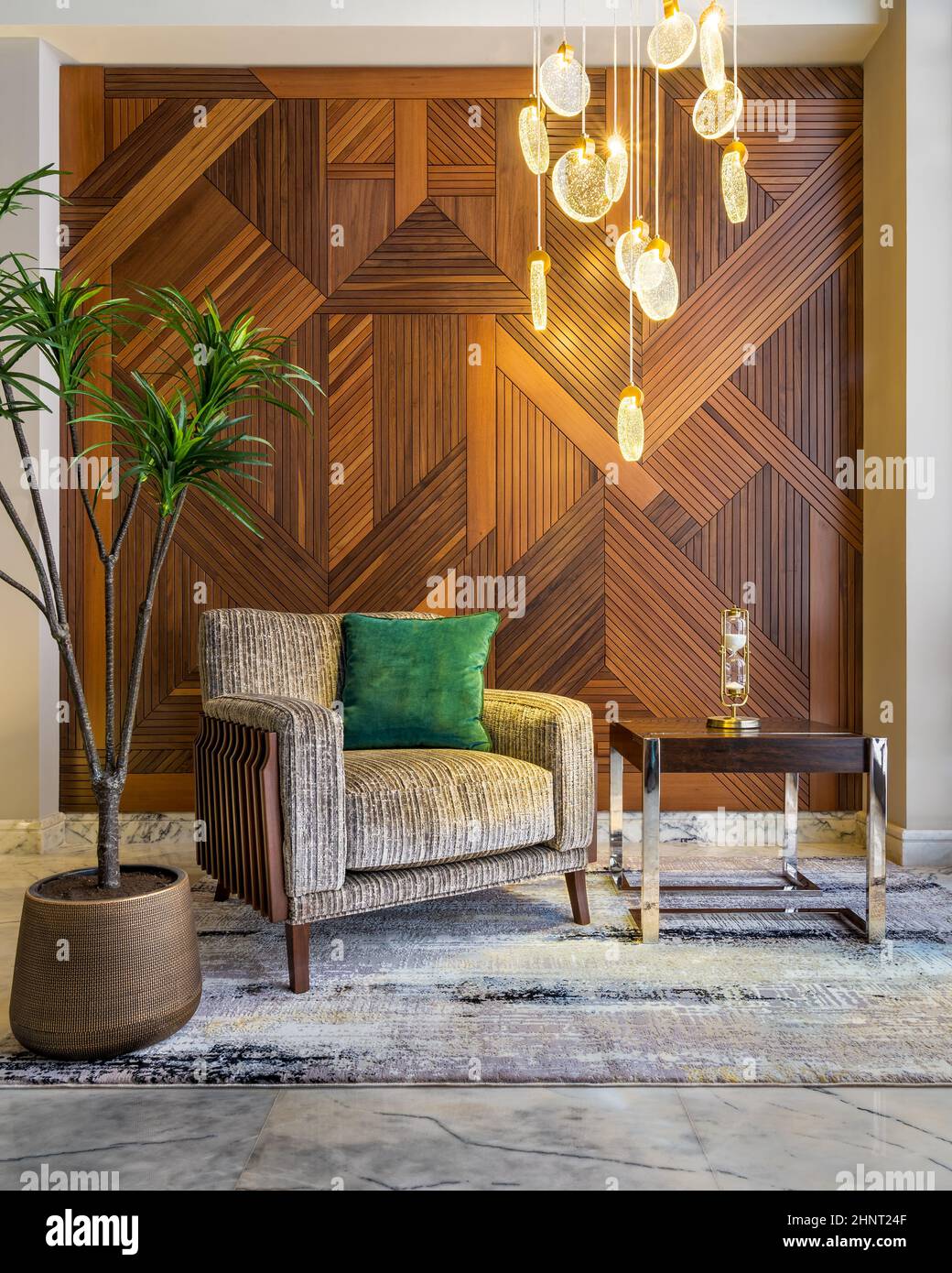 Moderner beigefarbener Sessel, kleiner Couchtisch, Blumentopf, moderner großer Glasleuchter und dekorierte Holzverkleidungen an der Wand Stockfoto