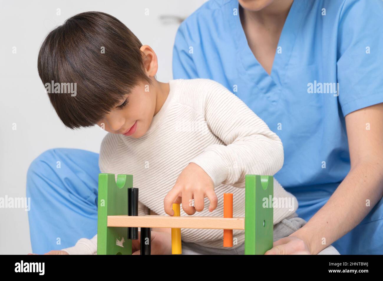 Niedliches Kind mit Behinderung spielt mit Spielzeug, während es von einem Physiotherapeuten im Reha-Krankenhaus unterstützt wird. Stockfoto